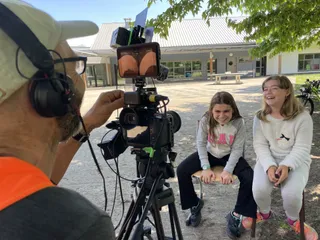 Marc filme deux collegiennes dans la cours de leur école. Elles sont asises et joyeuses en attendant de débuter le tournage. Marc règle le cadrage.