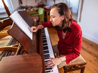 Paule Cornet, chemise rouge, est assise devant son piano en bois. Elle compose d'une main tout en notant les notes sur un carnet.